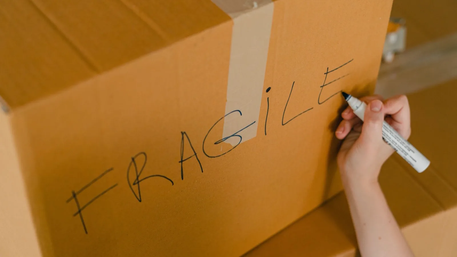 From above of crop unrecognizable person writing with marker on cardboard box with fragile products before relocation