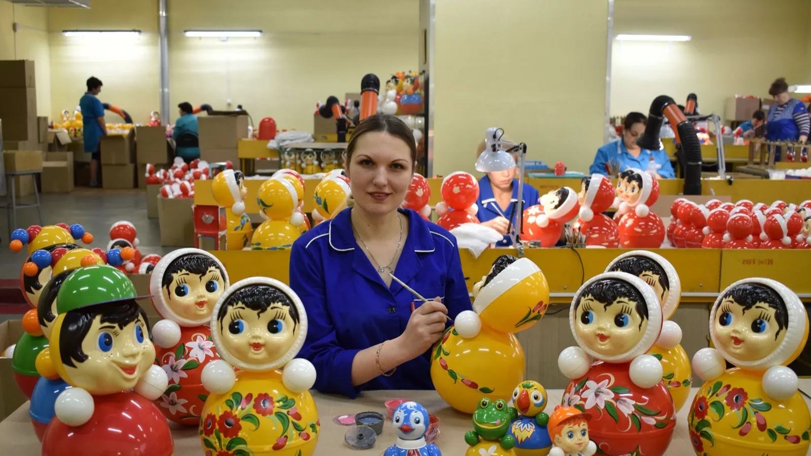 A woman in a toy factory carefully painting traditional colorful toys, surrounded by various dolls.