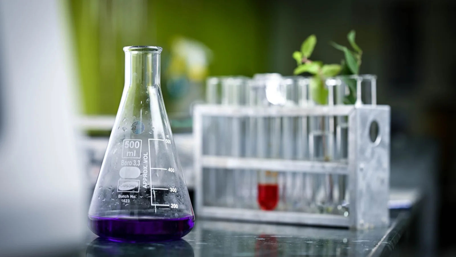 Close-up of a lab flask with purple liquid and test tubes in a scientific setting.
